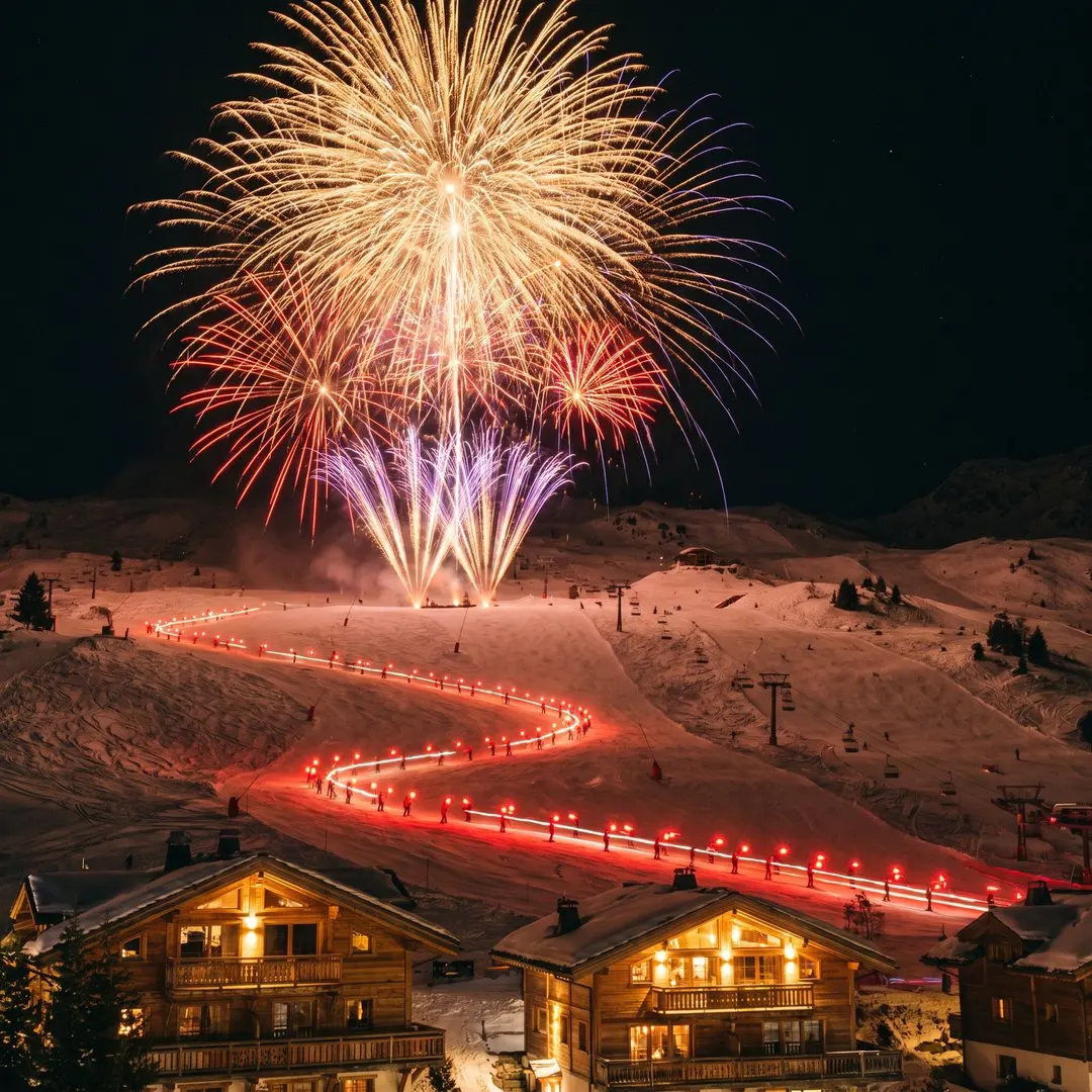 Fireworks bursting in gold, red and violet above the snowy slopes of Courchevel at night, a line of skiers carrying red torches descending the piste below, chalets lit warmly in the foreground, crisp clear sky