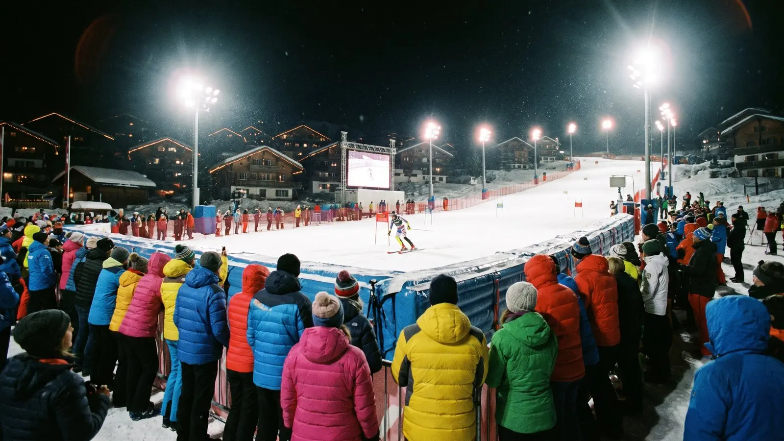Spectators in ski jackets along the barrier of a floodlit slalom finish area in Courchevel 1850 at night, a racer crossing the line under bright lights.